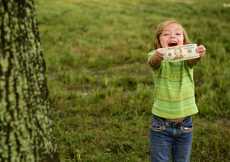 a young, excited child with blonde hair holds up a twenty dollar bill in the middle of a grassy field