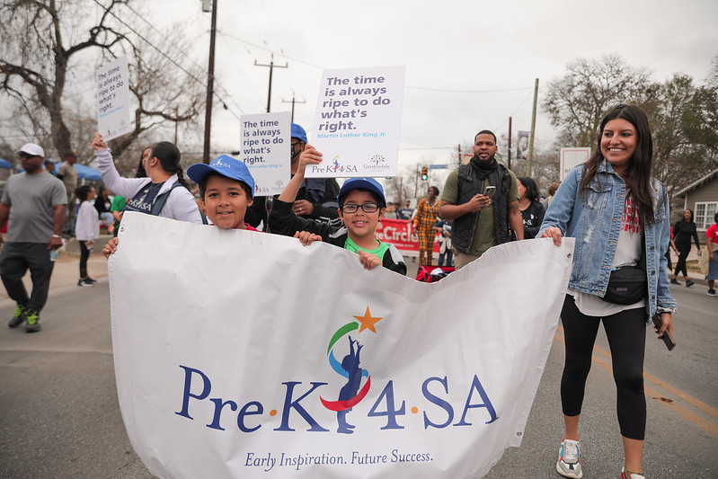 children and adults march down the road on Martin Luther King Day, while holding a banner that shows the Pre-K 4 SA logo