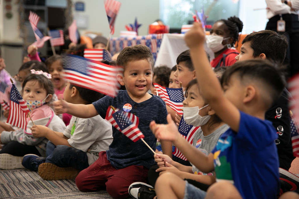 a group of young children sit on the floor and wave small American flags