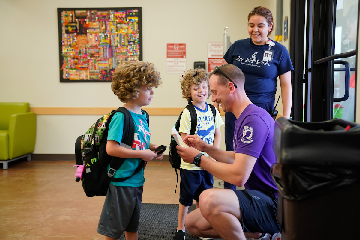 A Pre-K 4 SA staff member, parent, and two children greet each other at the door and smile.