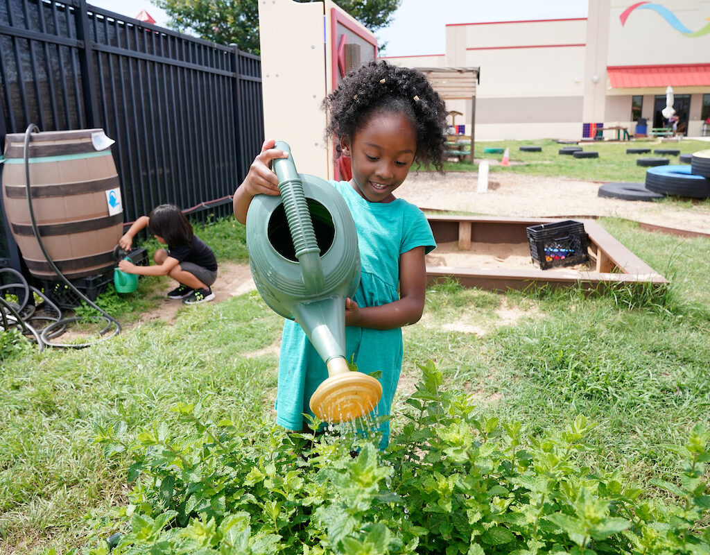 a young girl waters plants in a garden oustide a Pre-K 4 SA center