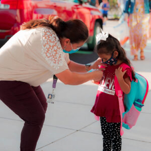 An adult leans over at a pre-k drop off to help a young child put on their name tag