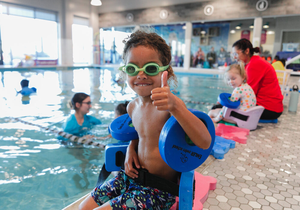 a young boy wearing swimming gear gives a thumbs up while sitting next to a pool