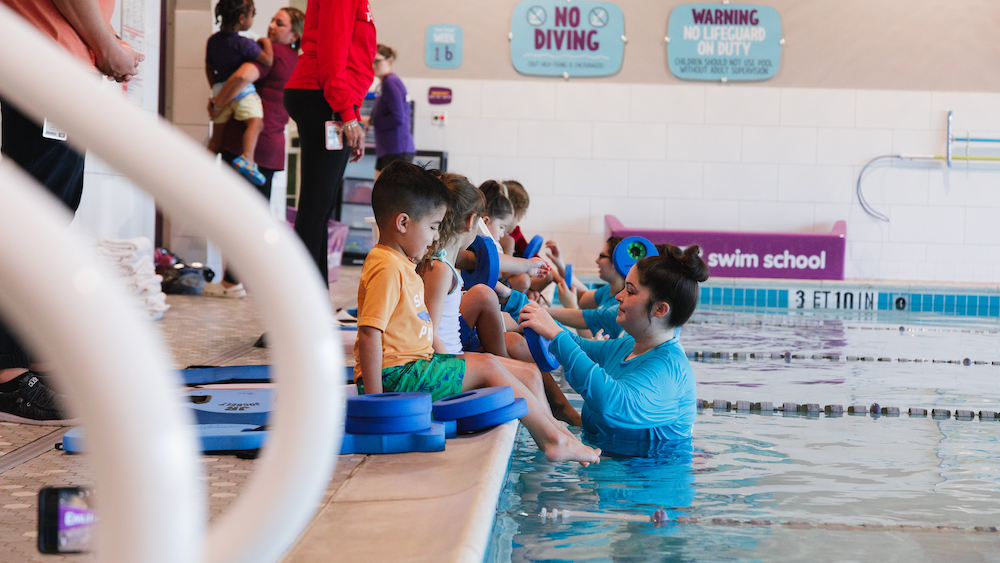 young children sit along the edge of a pool during a swimming lesson