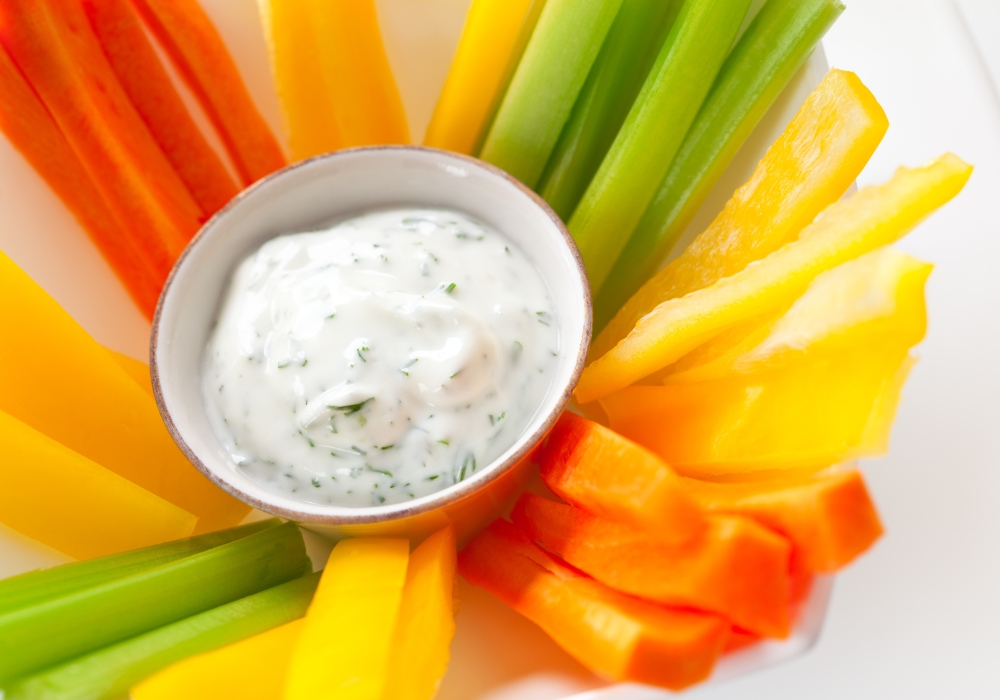 An array of carrot, celery, and bell pepper sticks around a small cup of dill veggie dip.