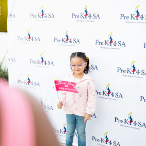 A young child holds a sign that says "first day of Pre-K" in front of a Pre-K 4 SA step-and-repeat.