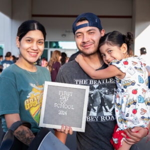 A father holds up his toddler and a sign that says "First Day of School"