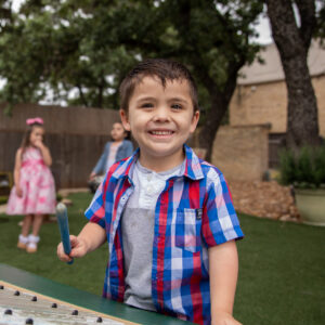 A child in a plaid shirt holds a drumstick and plays music in a garden at Pre-K 4 SA