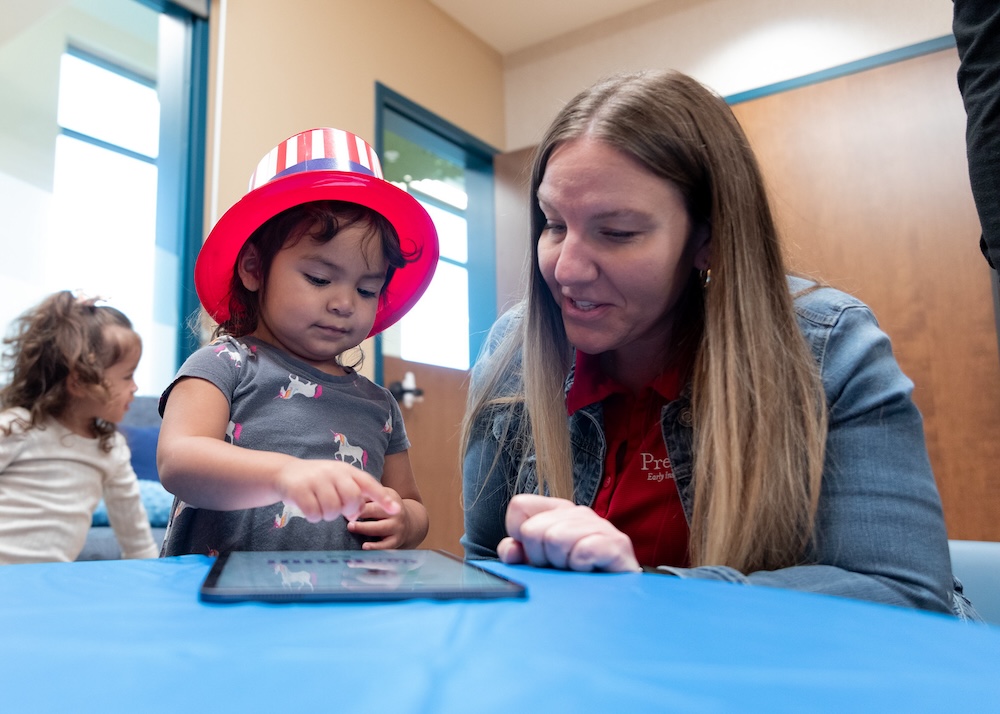 Bright-eyed preschool girl wearing a festive hat explores an educational app on a tablet with her teacher at Pre-K 4 SA.