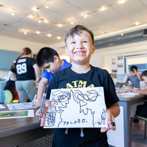 a young boy smiles and holds up a drawing