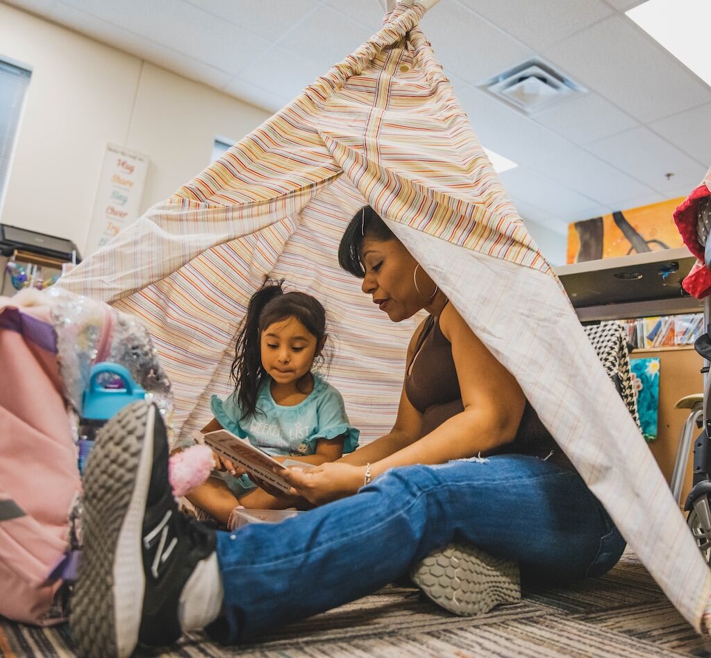 Pre-K 4 SA Partners With H-E-B Read 3 to Promote Reading at Home Adult and child reading inside a makeshift tent in a classroom setting.