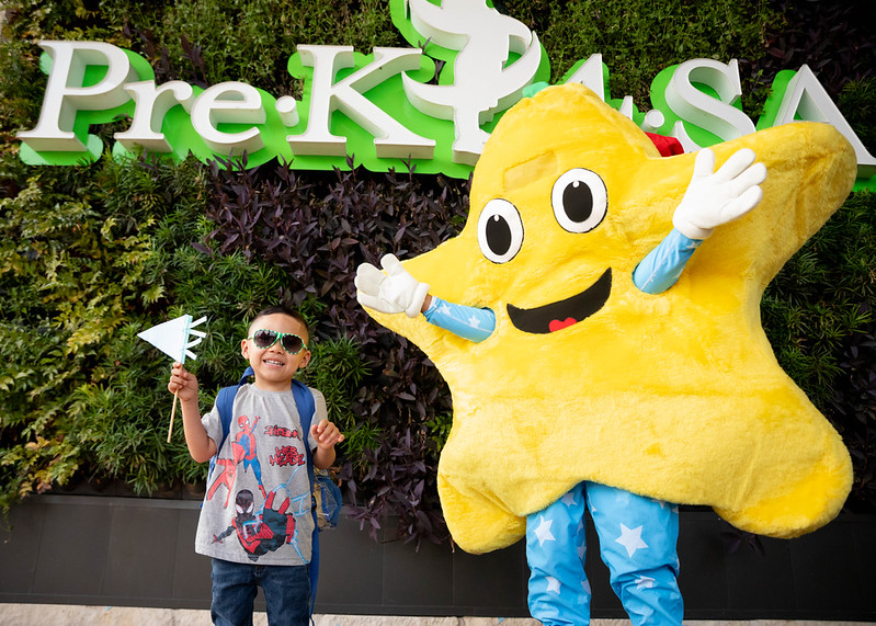 pre-k 4 sa child with rockstar at south Child with star-shaped flag standing next to Pre-K 4 SA mascot outside school.