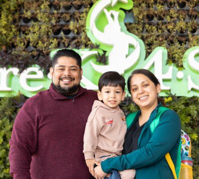 A smiling family of three in front of the Pre-K 4 SA logo at an outdoor event.