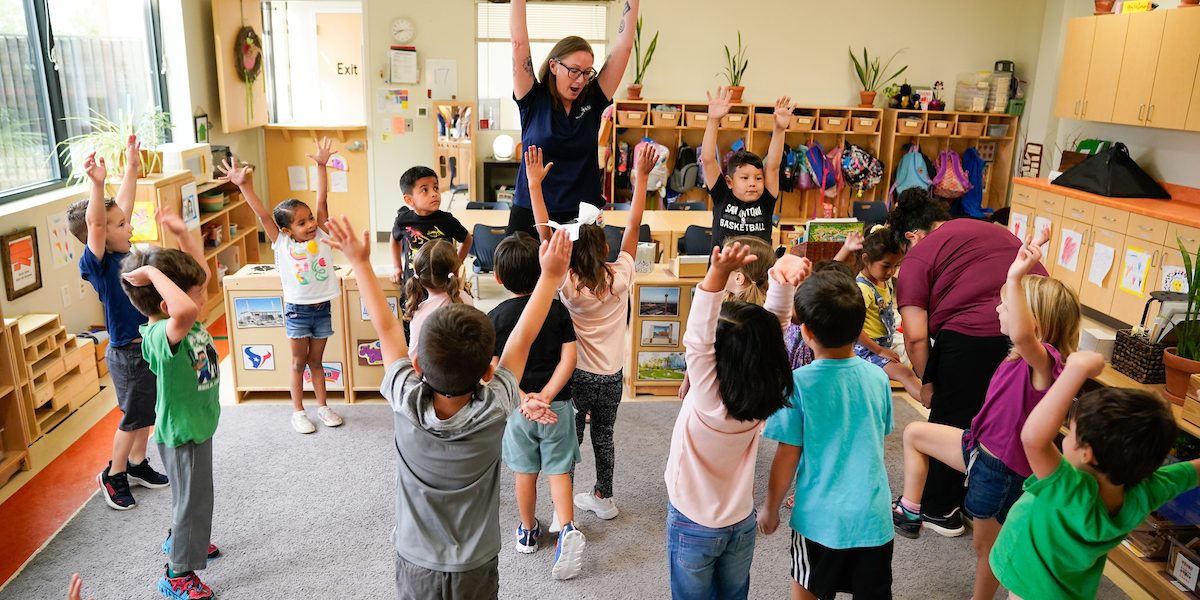 a group of young children and an educator reach for the sky in a classroom