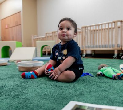 A toddler sitting on a green carpet in a preschool classroom with toys and books.