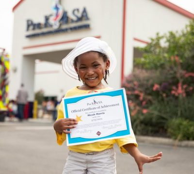 Young girl holding certificate outside Pre-K 4 SA school building.